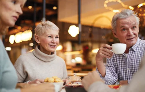 Three people are sitting at a table, enjoying a meal together. They are smiling and having a good time. A man is holding a cup of coffee, and there are several donuts on the table. The scene appears to be a casual gathering of friends or family. AI generated content